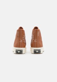 Pár hnědých vysokých tenisek Converse Chuck Taylor zobrazených zezadu na jednoduchém bílém pozadí.