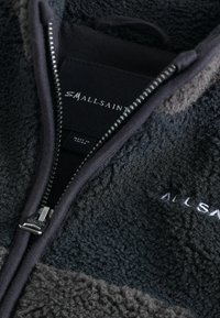 Juodai ir pilkai dryžuota fliso striukė su užtrauktuku, pasižyminti tekstūruotu audiniu, švariomis linijomis ir prekės ženklo etikete su užrašu "SMALLSAINT."