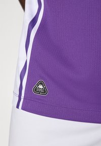 Maillot de sport violet avec un tissu texturé, des accents blancs et un logo noir en forme de triangle portant la mention "KOMBAT SYSTEM" près de l'ourlet.