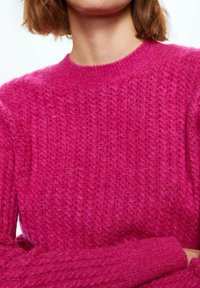 Vrouw in een felroze, getextureerde gebreide trui met lange mouwen en een ronde hals, met de armen gekruist, tegen een effen achtergrond.