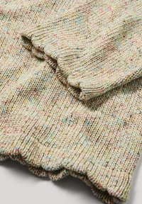 Gros plan sur un pull en maille beige plié avec des taches multicolores, montrant des poignets et un ourlet côtelés sur un fond blanc.