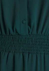 Tissu texturé vert foncé avec un bouton recouvert et un large détail de ceinture élastique froncée.