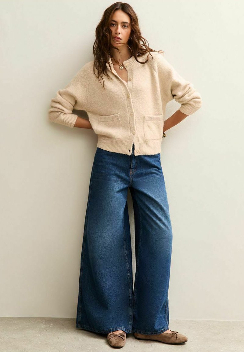 Cárdigan de punto beige con un corte relajado, que presenta un cierre de botones y dos bolsillos frontales. Combinado con unos vaqueros de denim azul de pierna ancha y unos zapatos planos marrones.