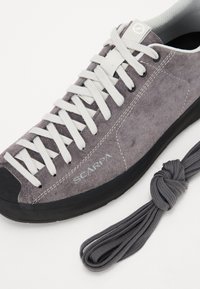 Graue Wildledersneaker mit weißen Schnürsenkeln, schwarzer Gummisohle und Logodetail. Enthält einen grauen geknoteten Schnürsenkel neben dem Schuh.