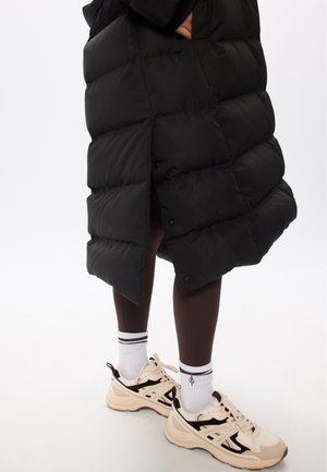 Giacca a vento nera con design trapuntato, caratterizzata da una fessura laterale. Indossata sopra leggings marroni e scarpe da ginnastica beige con dettagli neri.