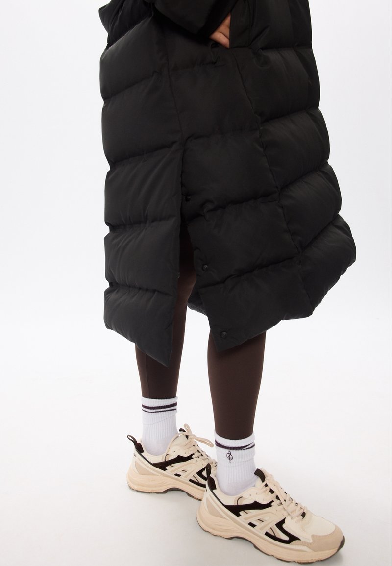 Manteau matelassé noir avec un design en piqué, doté d'une fente latérale. Porté sur des leggings marron et des chaussures de sport beiges avec des accents noirs.