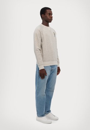 Sudadera gris claro con tejido texturizado, puños y bajo acanalados. Presenta un pequeño diseño bordado colorido en el pecho. Lleva puesta con jeans azules y zapatillas blancas.