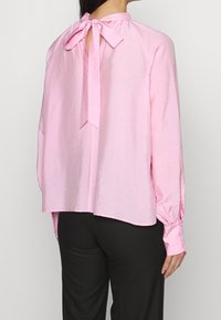 Blusa rosa con una vestibilità rilassata, lacci sullo scollo posteriore, maniche lunghe con dettagli a bottone, texture liscia e orlo curvo.