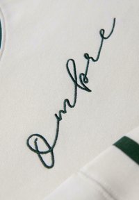 Tejido blanco con bordado cursivo en verde oscuro que dice "Onshore" y una franja verde a juego en un borde doblado.