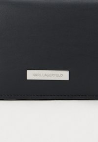 Černý kožený povrch s malou obdélníkovou stříbrnou destičkou, na které je vyryto "KARL LAGERFELD" velkými písmeny.