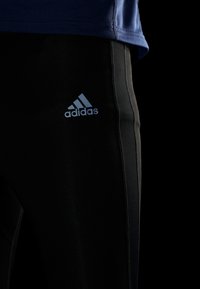 Pantaloni sportivi neri con una texture liscia e logo Adidas blu. Presentano una vestibilità affusolata e strisce laterali grigie in contrasto.