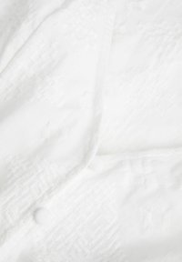 Tela blanca con sutiles patrones en relieve y botones circulares. Textura suave; el diseño presenta formas geométricas y un intrincado trabajo de costura.