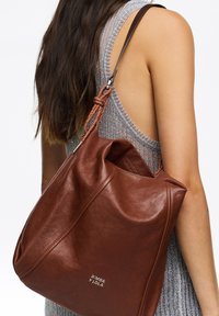 Rjava usnjena hobo torba s sproščenim dizajnom, kateri daje poudarek pletena naramnica in kovinski logotip. Gladka textura in prostoren odprtina.