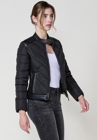 Giacca trapuntata nera con design aderente, dotata di un alto colletto, dettagli in pelle a coste e zip laterali. Indossata sopra una maglietta bianca.