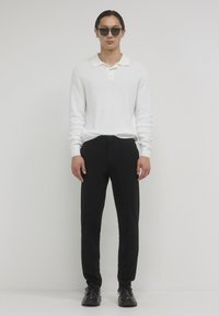 Mann mit schwarzen Sonnenbrillen, weißem Langarm-Poloshirt, schwarzen Hosen und schwarzen Schuhen, der vor einem schlichten hellen Hintergrund steht.