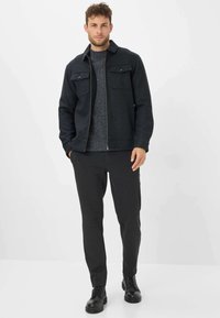 Donkere wollen jas met twee borstzakken, grijze gebreide trui en donkere broek. Zwarte veterlaarsjes maken de outfit compleet.