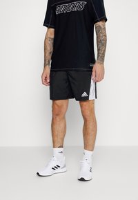Camiseta atlética negra con el audaz logo blanco de "Adidas", combinada con pantalones cortos negros que tienen un acento blanco. Zapatillas deportivas blancas completan el atuendo.