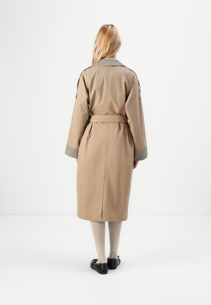 Femme debout de dos, portant un trench-coat beige ceinturé avec un col et des poignets à carreaux, des collants beiges et des ballerines noires sur fond blanc.