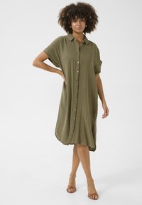 Rochie verde oliv, cu nasturi, maneci scurte, croi lejer, material texturat, până la genunchi. Purtați cu sandale cu barete, design minimalist.