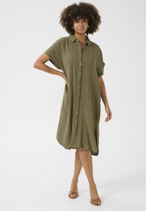 Abito button-up verde oliva, maniche corte, vestibilità ampia, tessuto testurizzato, lunghezza al ginocchio. Indossato con sandali con cinturini, design minimalista.