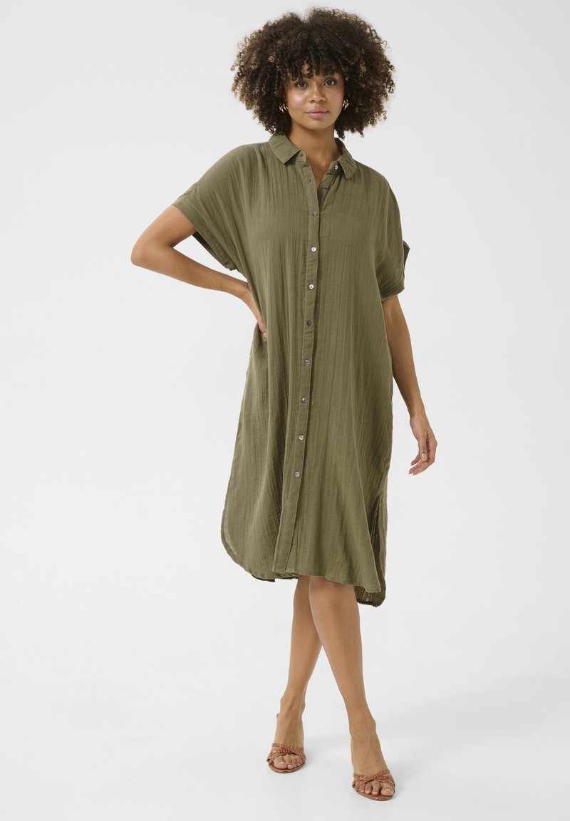 Rochie verde oliv, cu nasturi, maneci scurte, croi lejer, material texturat, până la genunchi. Purtați cu sandale cu barete, design minimalist.