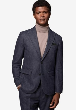 Veste de costume texturée bleu marine avec revers en pointe et deux poches avant, associée à un col roulé beige clair, agrémentée d'un accent de pochette.