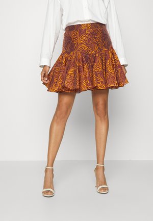 Woman wears flared skirt with orange and brown leaf pattern, paired with white buttoned shirt and white ankle-strap heels.