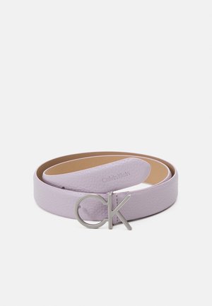 Liliowy pasek skórzany zwinięty z srebrną metalową klamrą z logo "CK" oraz wytłoczonym napisem Calvin Klein blisko końca na białym tle.