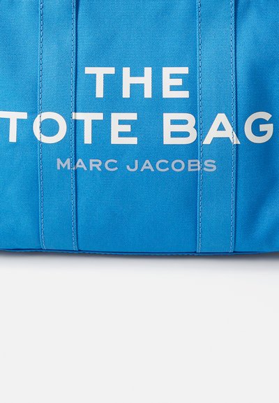 Sac fourre-tout en toile bleue avec un texte blanc audacieux : "LE SAC FOURRE-TOUT" et "MARC JACOBS" dans une police moderne et épurée. Finition texturée avec des accents cousus.