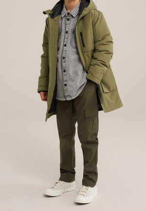 Enfant portant un manteau à capuche vert olive, une chemise grise boutonnée, un pantalon cargo olive et des baskets blanches, debout sur un fond neutre.