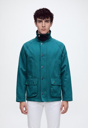 Un homme porte une veste bleu canard boutonnée sur le devant, à col montant et grandes poches à rabat, assortie à un pantalon blanc sur fond uni.