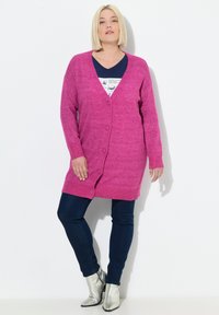 Cardigan lungo rosa con trama a maglia, caratterizzato da scollo a V e cinque bottoni. Abbinato a una maglietta blu navy, jeans scuri e stivaletti argento.