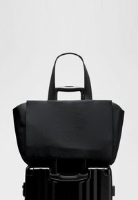 Melnas auduma soma, piestiprināta pie melna rullīša koferi ar izvilktu rokturi uz vienkārša gaiša fona.