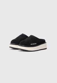 Črne semiš clogs s belo platformo, ki ima teksturirano črno podlago in oznako "LOVE MOSCHINO" na strani.