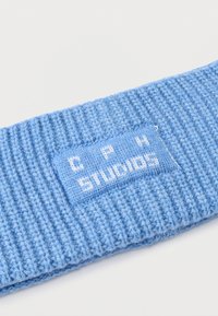 Niebieska dziana opaska na głowę o prążkowanej fakturze, z naszytą prostokątną metką z napisem "C P H STUDIOS" białymi literami.