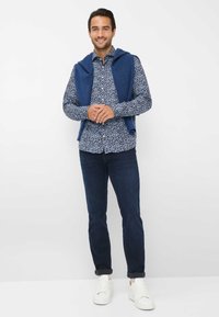 Camicia a fantasia floreale blu, maglione navy appoggiato sulle spalle, jeans scuri e sneakers bianche. Tessuto liscio, design con bottoni, look casual.