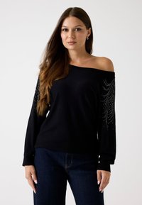 Schwarzer, schulterfreier Pullover mit langen Ärmeln, verziert mit silbernen Verzierungen. Weiche Textur, leichtes Material, kombiniert mit dunklen Jeans.