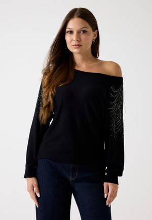 Zwarte off-the-shoulder trui met lange mouwen versierd met zilveren embellissementen. Zachte textuur, lichtgewicht materiaal, gecombineerd met donkere spijkerbroek.