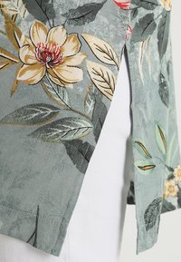 Floral-patterned fabric in shades of green, gold, and beige; features smooth texture, short sleeves, and side split detailing.