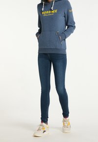 Osoba v modré mikině "NORDSEE", tmavých skinny jeanech a bílých teniskách s žlutými a růžovými detaily, stojící se založenýma rukama v kapsách.