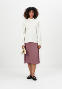 Donna che indossa una blusa bianca a maniche lunghe con texture, una gonna midi mauve, ballerine nere e tiene una clutch intrecciata beige contro uno sfondo semplice.