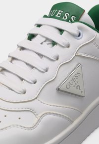 Baskets en cuir blanc avec lacets blancs, languette verte affichant le texte "GUESS" et logo métallique triangulaire Guess sur le panneau latéral.