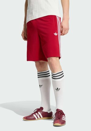 Červené šortky Adidas s bílými pruhy, doplněné bílými podkolenkami a červenými teniskami Adidas s bílými pruhy.