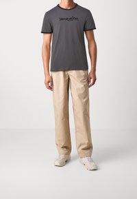 Camiseta gris de manga corta con logo gráfico negro, combinada con pantalones holgados beige y zapatillas de color claro.