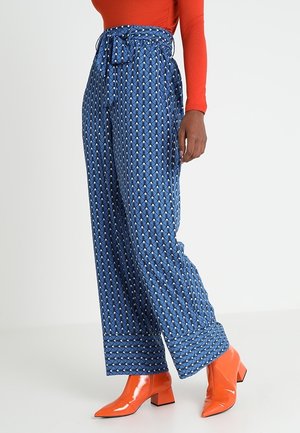 Frau trägt hoch taillierte, weit geschnittene blaue gemusterte Hose, ein rotes Langarmoberteil und glänzende orangefarbene Stiefeletten.