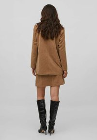 Hnědý kordový blazer a odpovídající sukně, doplněné černými vysokými botami. Texturovaná tkanina a upravený design s délkou k bokům.