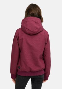 Bordeaux hettegenser med hette, teksturert stoff, elastiske mansjetter og midje. Har en logopatch på venstre underkant.