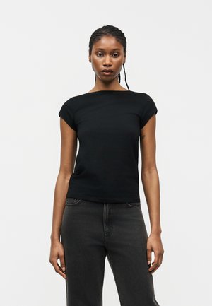 Νεαρή γυναίκα με πλεξούδες, φορώντας εφαρμοστό μαύρο κοντομάνικο τοπ και σκούρο γκρι τζιν, στέκεται μπροστά σε απλό λευκό φόντο.