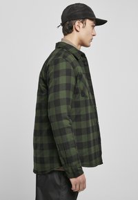 Zielono-czarna koszula w kratkę z zapinaniem na guziki, długimi rękawami i kieszenią na piersi. Nosi ją w połączeniu z czapką w kolorze czarnym, zakładając ją na beżową koszulę.