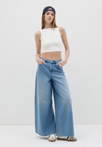 White cropped tank top, wide blue denim jeans with a high waistband, and white sneakers with brown accents. Model wears a dark cap.